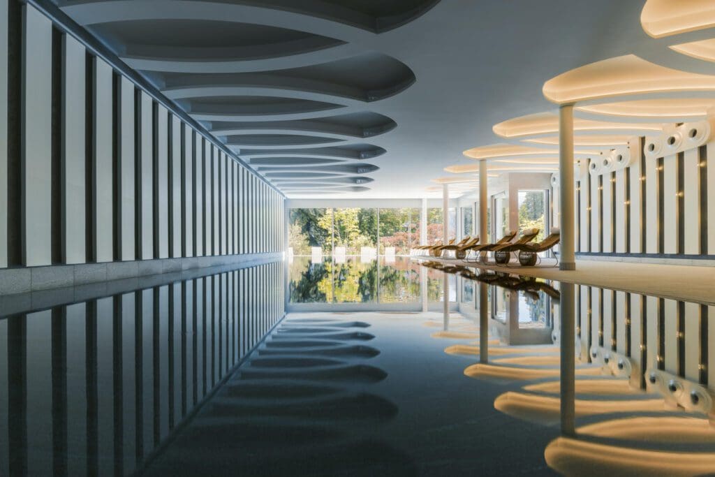 Indoor swimming pool with modern architectural design, lounge chairs on the right, large windows on the left, and ceiling lights reflected in the still water.