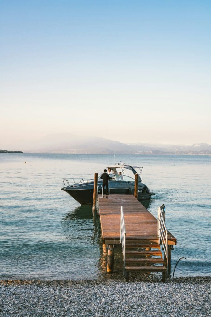 A black motorboat is moored at the end of a wooden pier on a calm body of water, with a person standing on board and mountains in the distance under a clear sky.