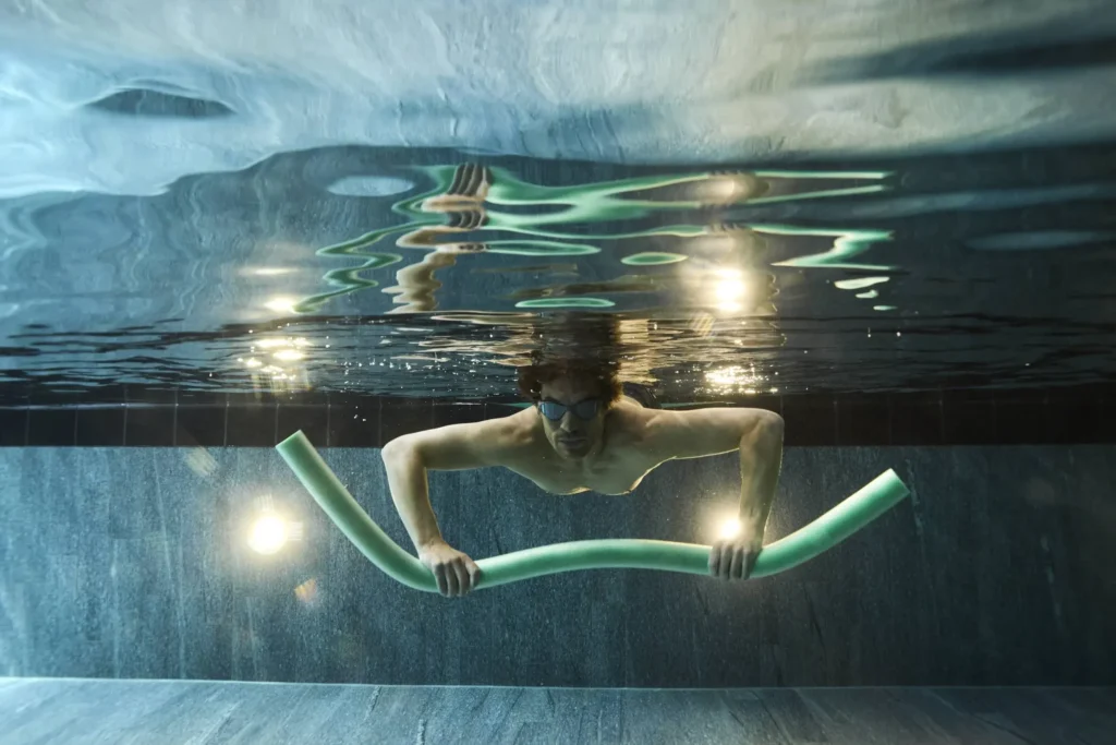 Person wearing goggles, submerged in a swimming pool, holds a green pool noodle horizontally with both hands under the water—focused like a strategist planning a PPC or Paid Search campaign.