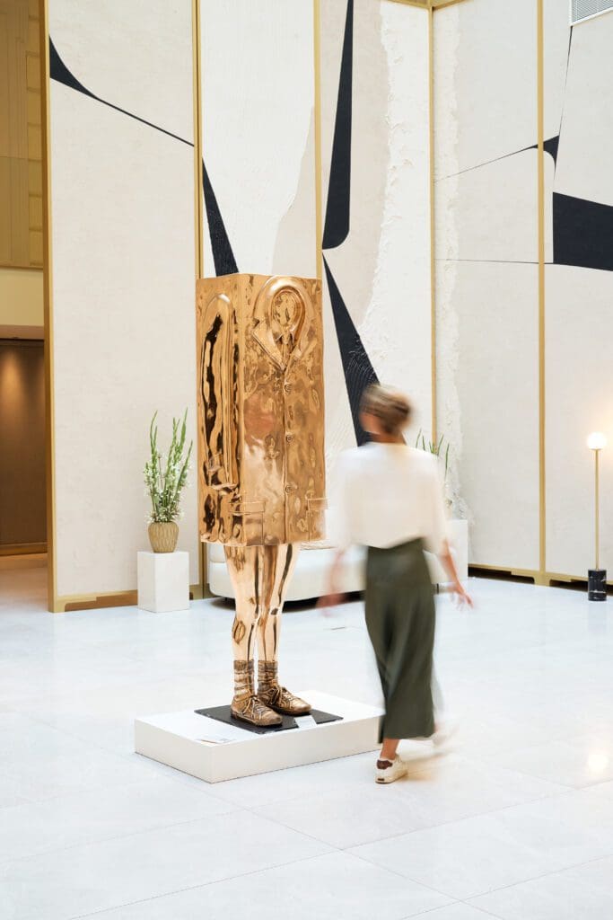 A person walks past a metallic sculpture of a human figure with a box covering its upper body, displayed on a white plinth in a modern, minimalist interior space.