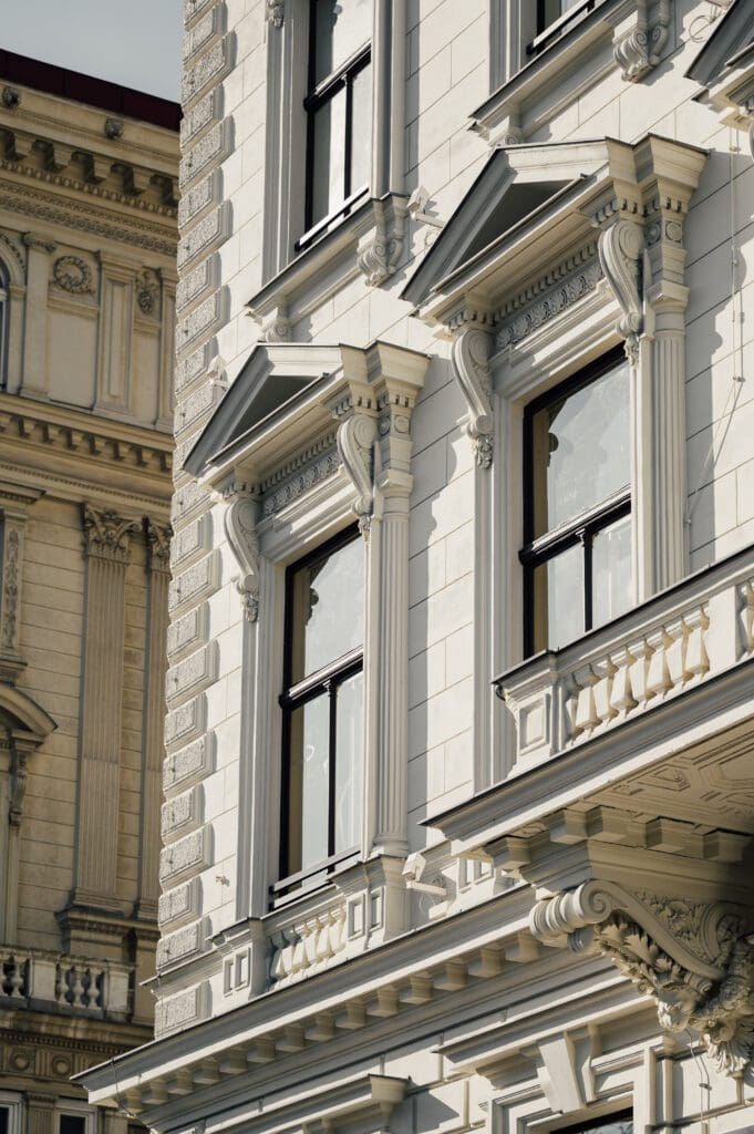 Close-up of ornate, classical-style building facade with decorative columns, window frames, and balconies; another similar building is partially visible in the background.