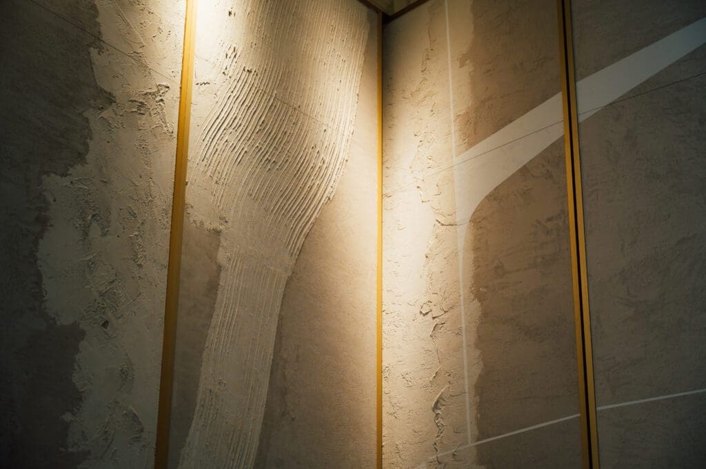 Textured beige wall panels with gold vertical trim are illuminated by soft, angled light, highlighting abstract patterns on the wall surface.