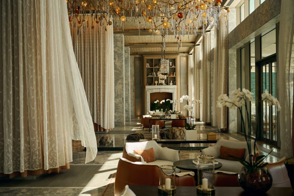 Elegant hotel lounge with marble floors, sheer curtains, modern seating, chandeliers, and large windows letting in natural light. Orchids and candles decorate the space.