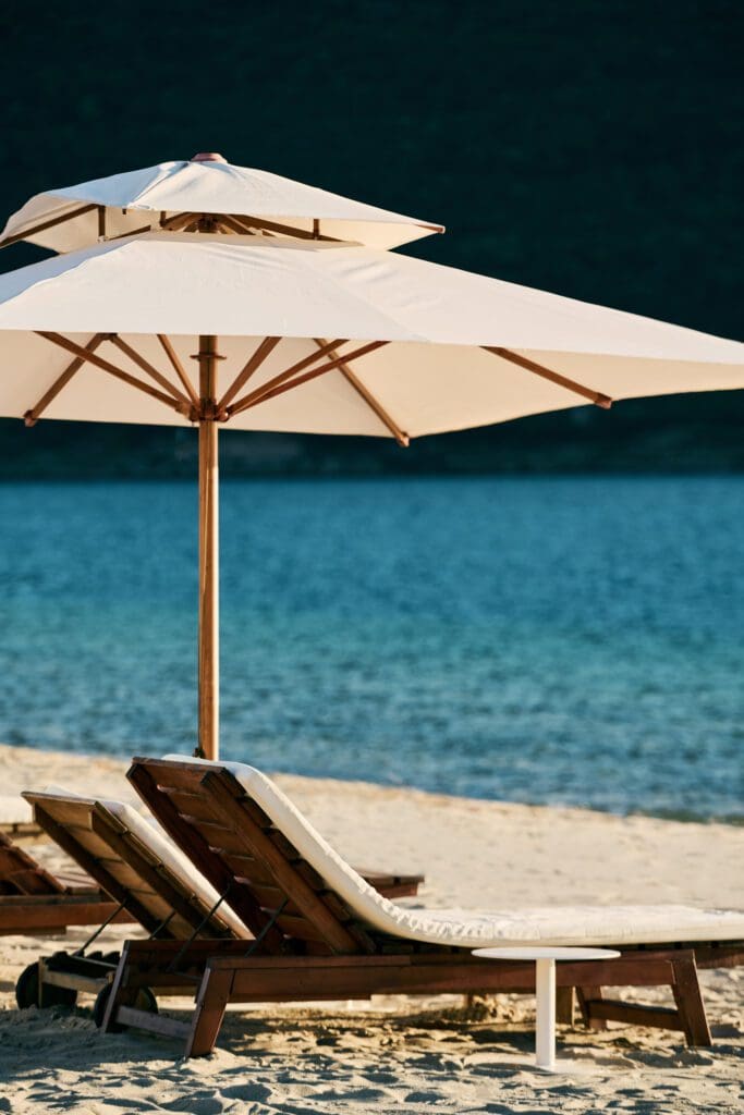 Two wooden loungers with white cushions sit under a large white parasol on a sandy beach near calm blue water.