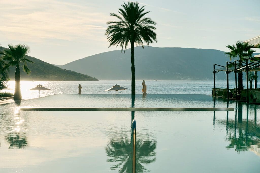 Infinity pool with palm trees overlooks calm sea and distant hills at sunset, with closed parasols and reflections on the water.