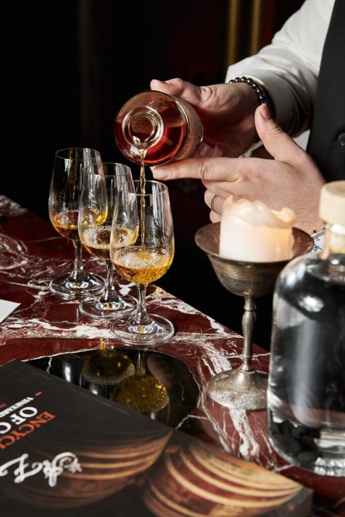 A person pours amber liquor into three wine glasses on a marble table next to a lit candle, a magazine, and a glass bottle.