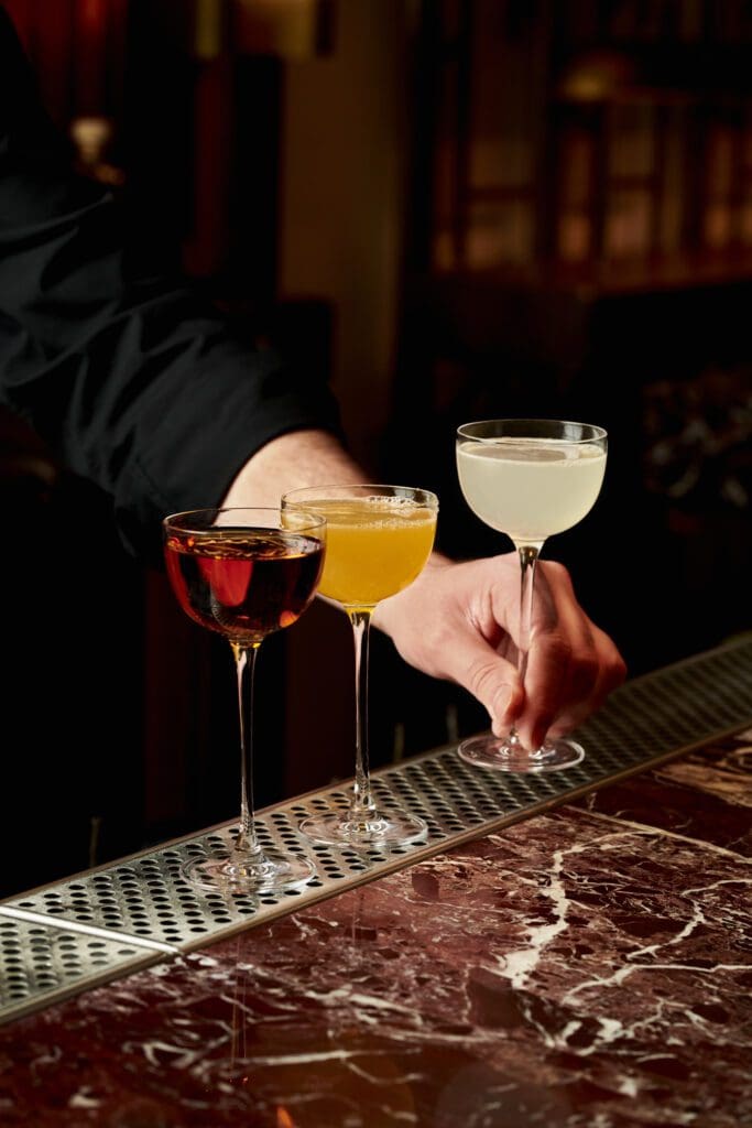 A person arranges three cocktails in different glasses on a marble bar counter: one dark red, one yellow, and one light yellow, with a metal bar mat underneath.