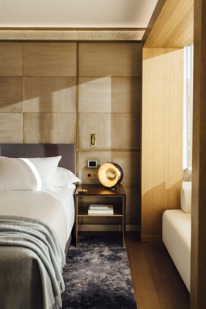 Modern hotel room with a neatly made bed, a bedside table holding a decorative lamp and books, natural wood accents, and sunlight streaming through a window.