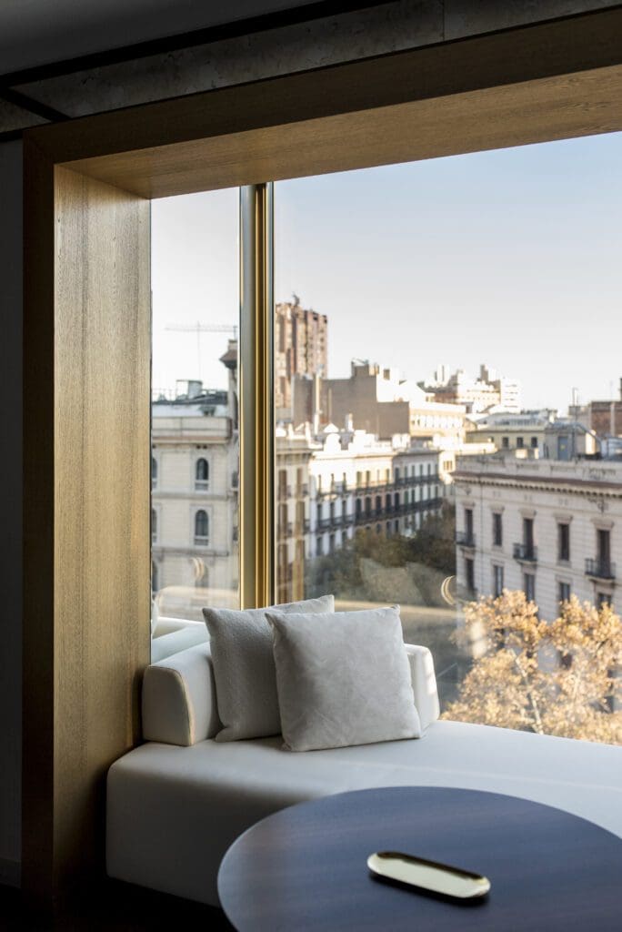 A modern window seat with white cushions overlooks historic city buildings and a tree-lined street, with a round table in the foreground.