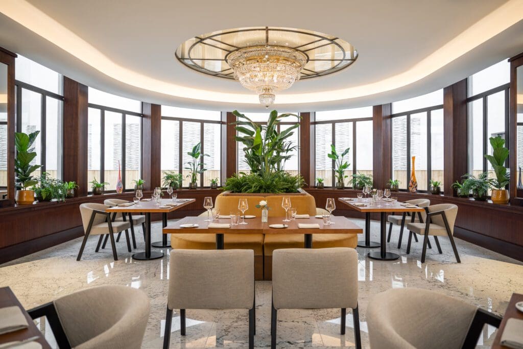 Modern restaurant dining area with round chandelier, large windows, green plants, beige chairs, and set tables on polished marble floor.