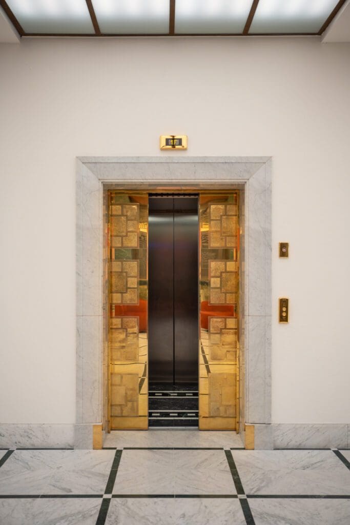 An open lift with gold and marble trim is centred in a white corridor with marble floor and walls; buttons and an indicator light are visible beside the door.
