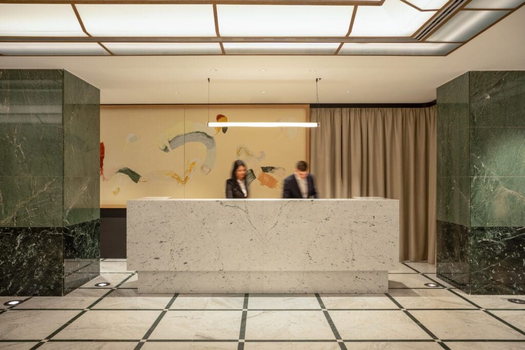 Two people stand behind a marble reception desk in a modern lobby with green marble walls, abstract art, and a ceiling light fitting.