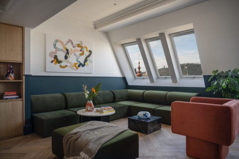 Modern living room with green corner sofa, coral armchair, round coffee table, abstract wall art, large windows, and a potted plant.