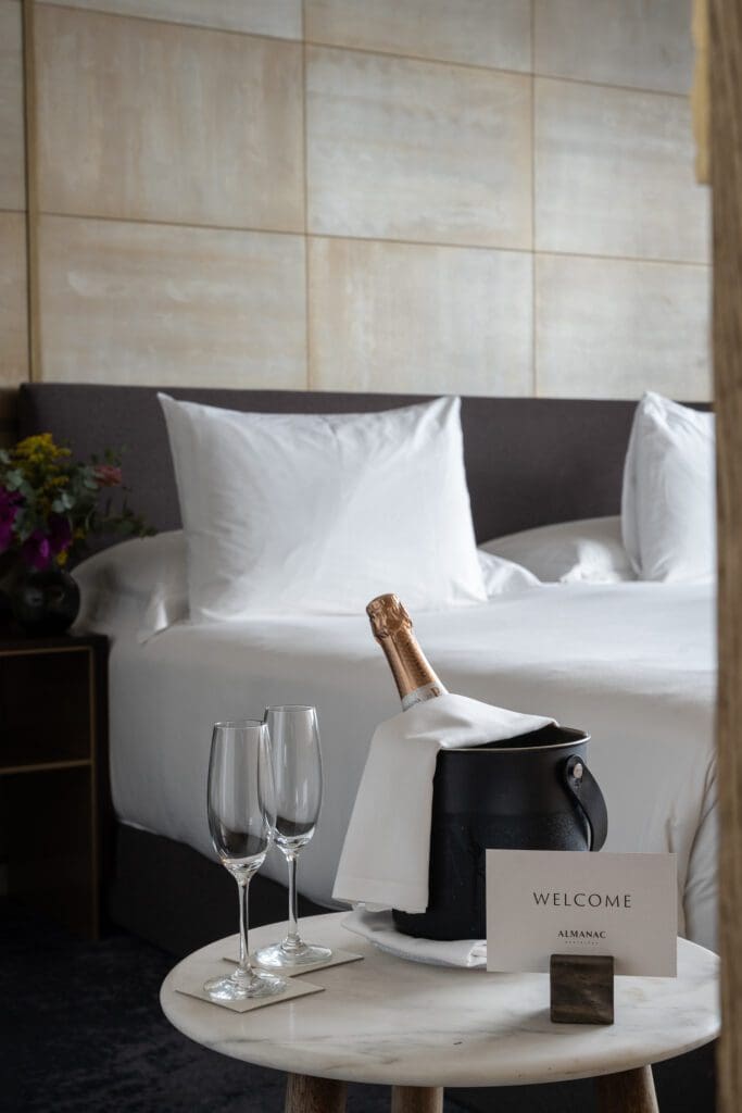 A hotel room with a neatly made bed, a bedside table with flowers, and a small table displaying a bottle of champagne in an ice bucket, two glasses, and a welcome card.