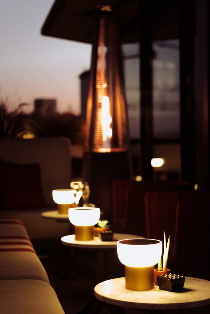 Three small round lamps glow on side tables beside a sofa on an outdoor patio at dusk, with a tall patio heater and blurred city buildings in the background.