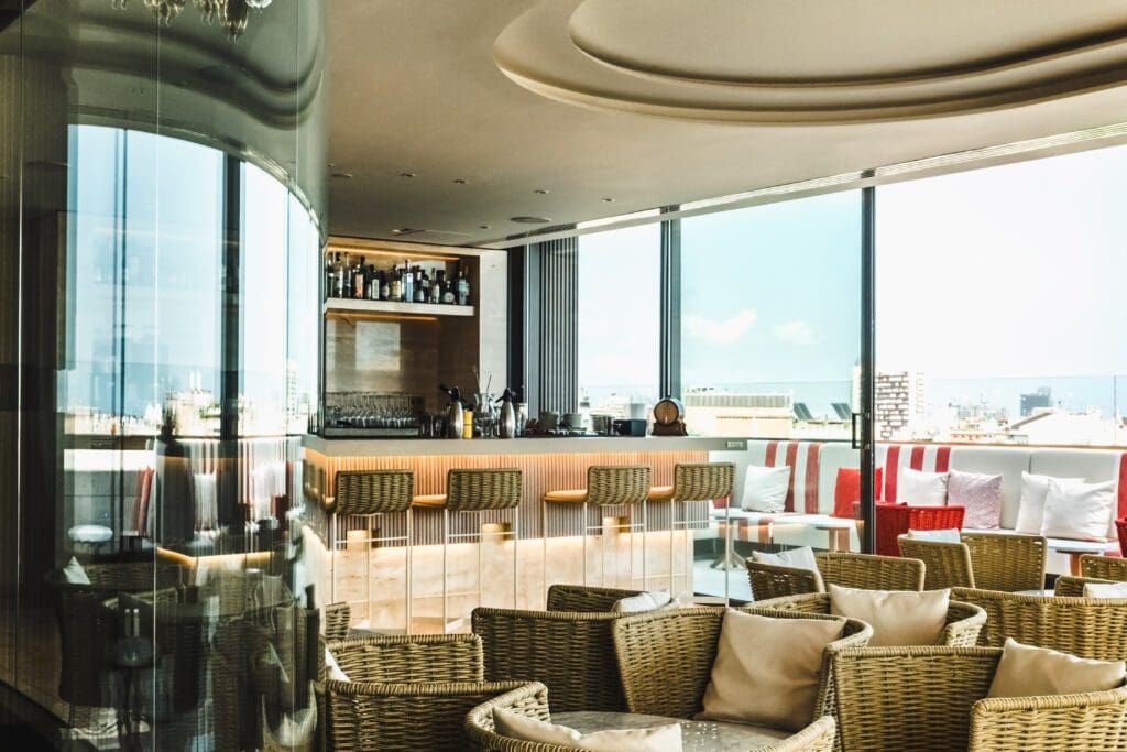 Modern rooftop bar with wicker chairs, a wooden bar counter, and large windows showing a city view under a bright sky.