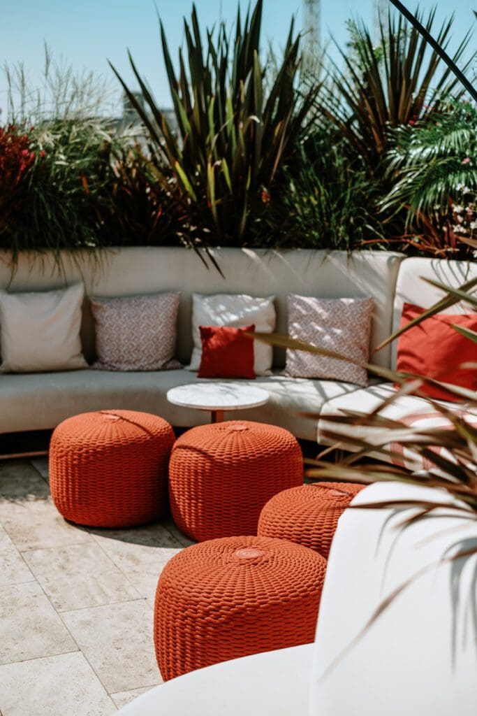 Outdoor patio area with a curved white sofa, assorted cushions, a small round table, and three orange woven pouffes, surrounded by green plants.