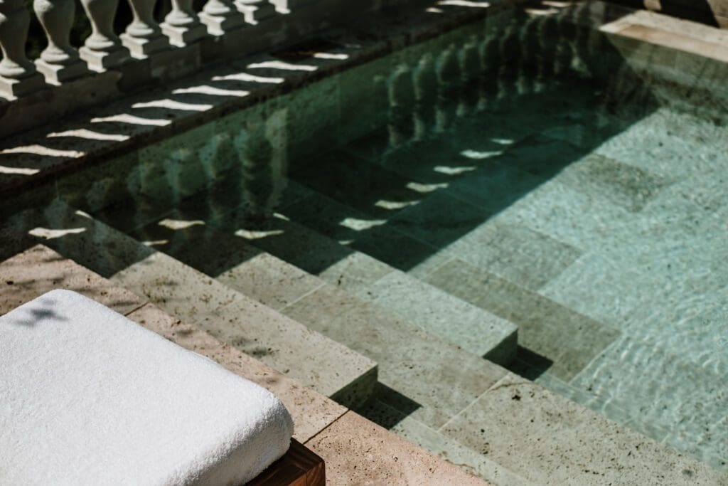 A white towel sits on a lounger beside a clear swimming pool with stone steps under bright sunlight.