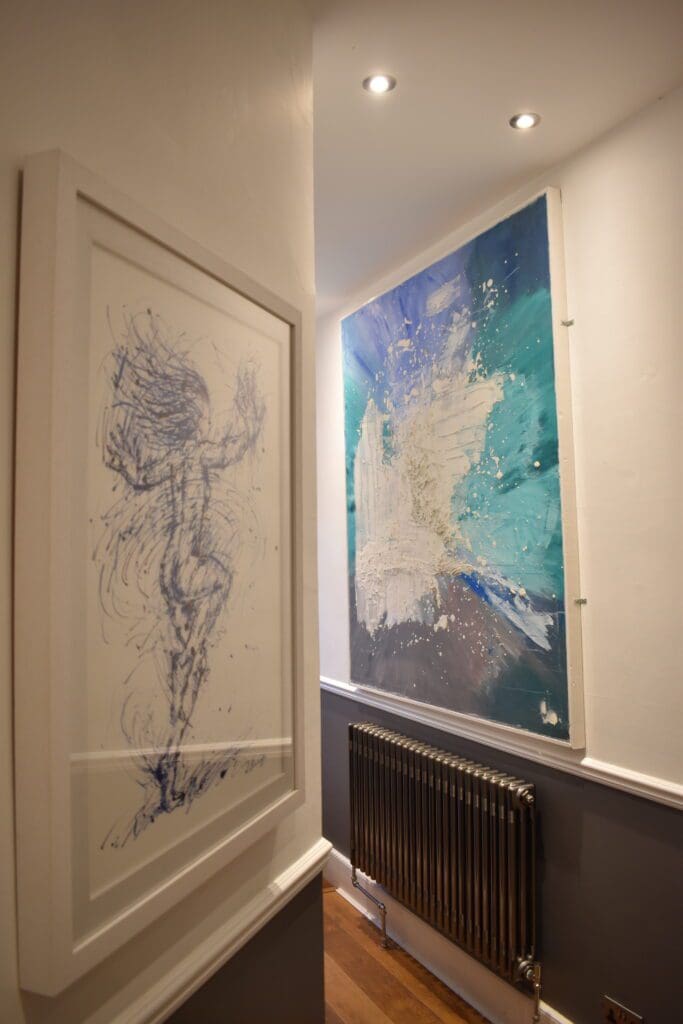 A corridor with two framed artworks on the walls, one featuring a blue abstract figure and the other displaying an abstract painting with blue and white tones above a radiator.