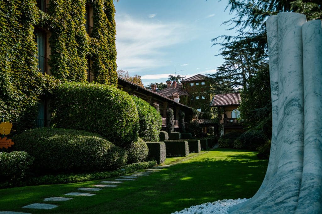 A lush garden with trimmed hedges and a stone path runs alongside a building covered in ivy, with a white abstract sculpture in the foreground.