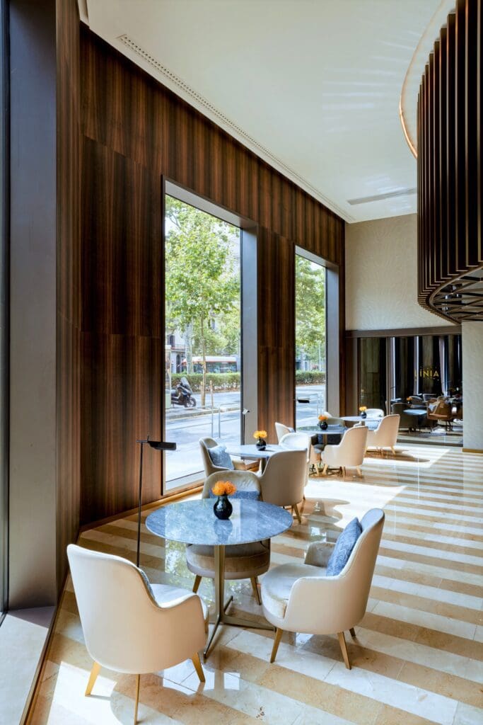 Modern café interior with round tables, white chairs, large floor-to-ceiling windows, wood panelling, striped flooring, and potted flowers on each table. Daylight streams in from outside.