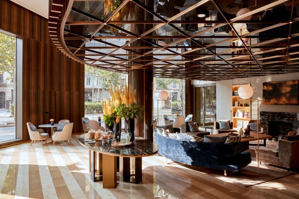 Modern hotel lobby with large windows, marble floors, blue seating, a black marble table with flowers, and a geometric mirrored ceiling. Natural light fills the space.