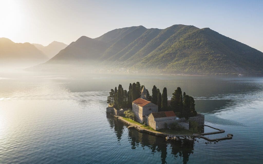 A small stone monastery with red roofs sits on an island surrounded by calm water, with tree-covered mountains in the background under a hazy sky.