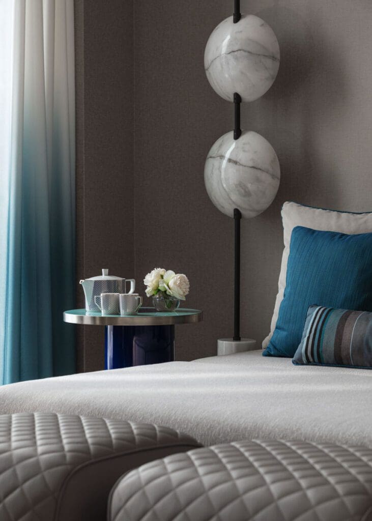 A neatly made bed with blue and white pillows, a bedside table holding a teapot set and flowers, and two round marble wall lights beside floor-length, gradient blue curtains.