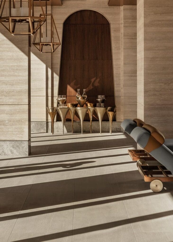 A modern interior with beige stone walls, a wooden accent, a gold console table with glassware, geometric pendant lights, and a cushioned lounge chair.