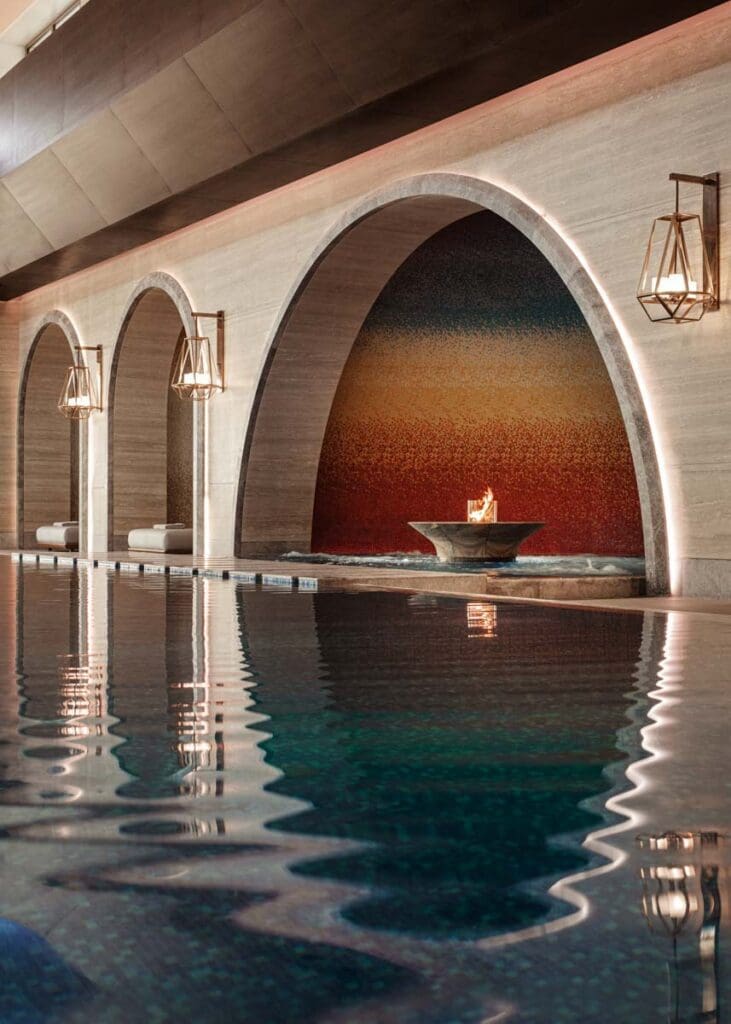 Indoor swimming pool with arched stone walls, wall-mounted lanterns, and a fire feature set against a colourful mosaic backdrop.