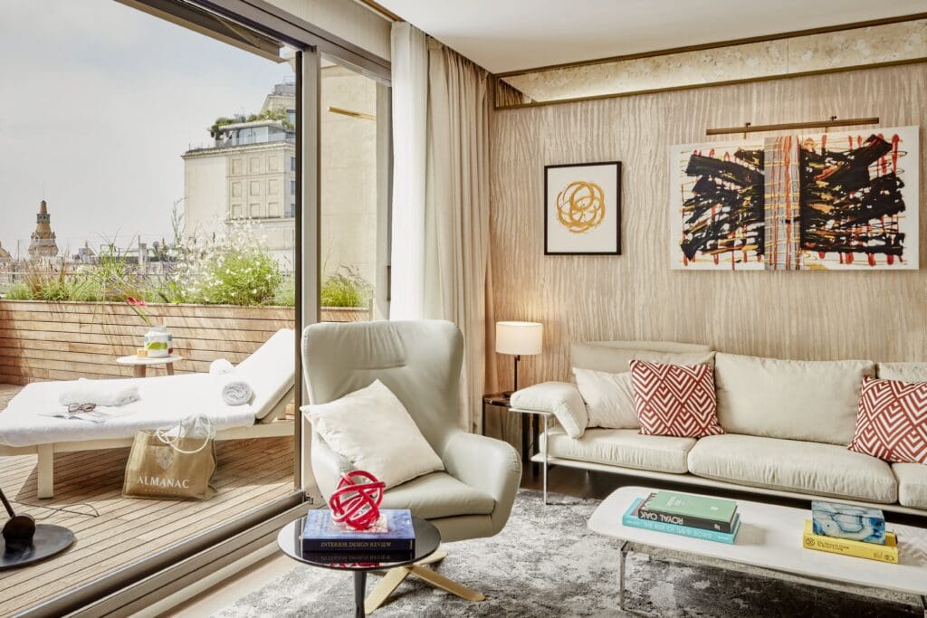 A modern living room with a white sofa, armchair, coffee table, and abstract art, opening onto a balcony with a lounger and city view.