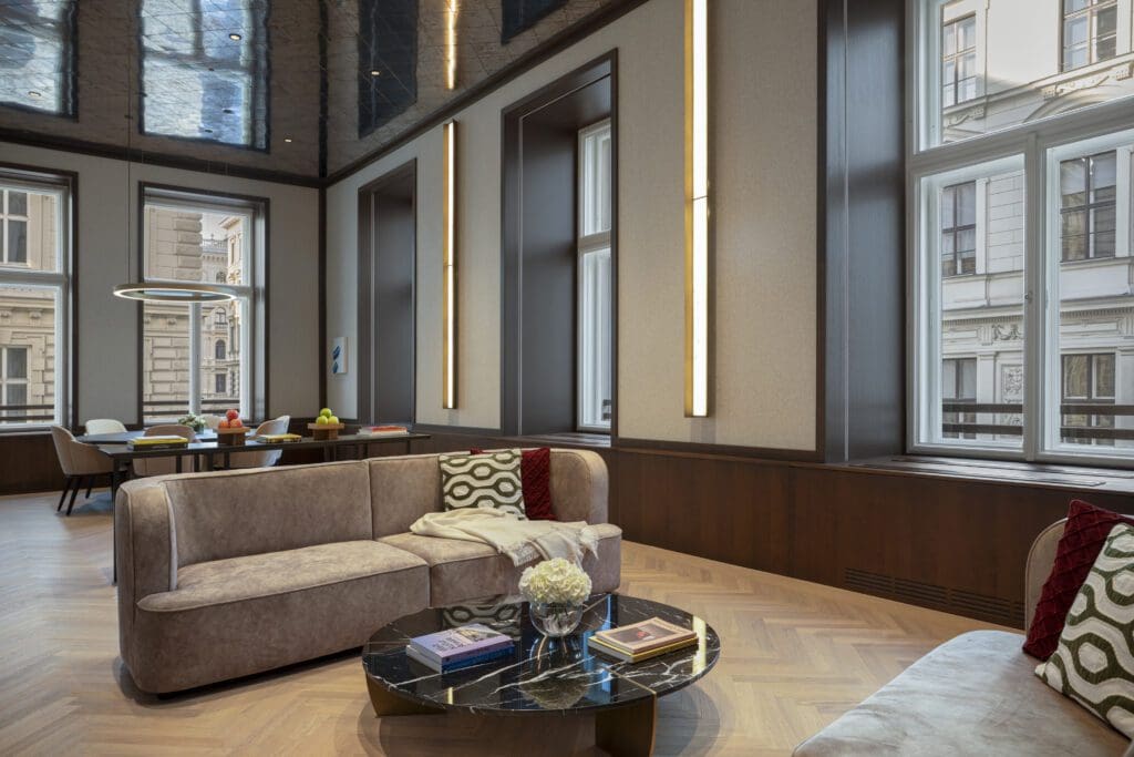 Modern living room with neutral-toned sofas, a black marble coffee table, books, and large windows letting in natural light. Dining area visible in the background.