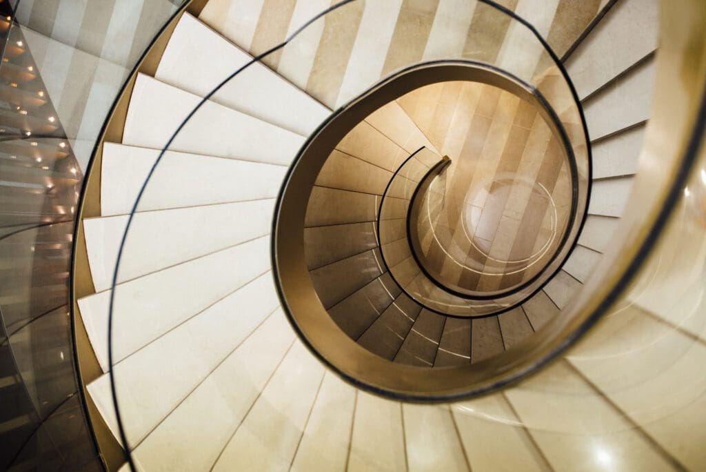 Top-down view of a modern spiral staircase with beige steps and a glass balustrade, creating a symmetrical, circular pattern.
