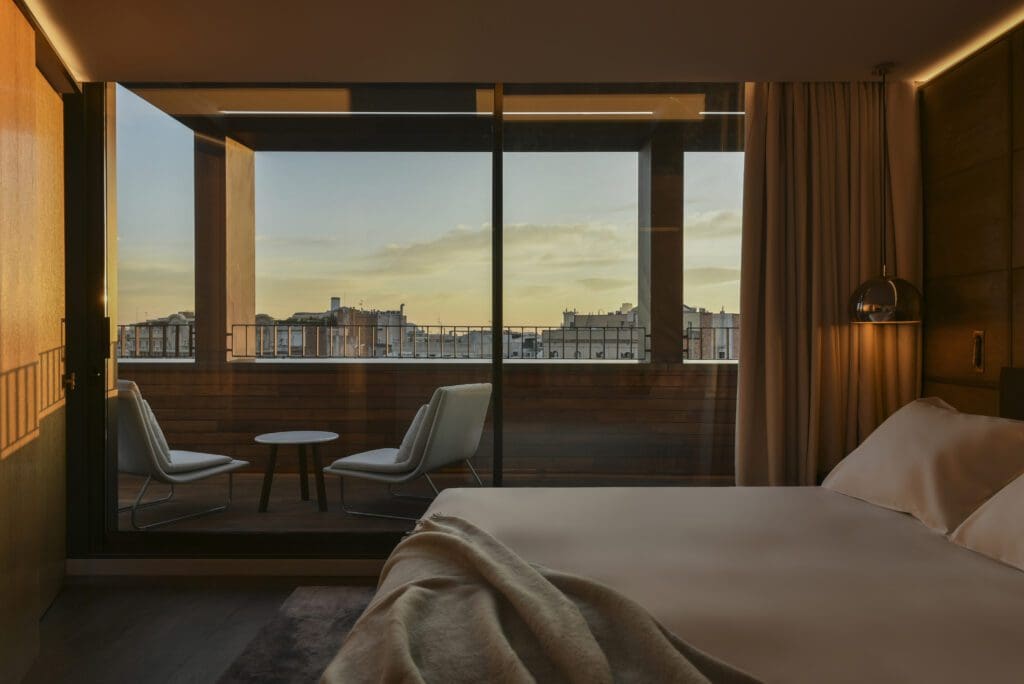 A modern hotel room with an unmade bed, floor-to-ceiling window, and a balcony featuring two chairs, a small table, and a cityscape view at sunset.