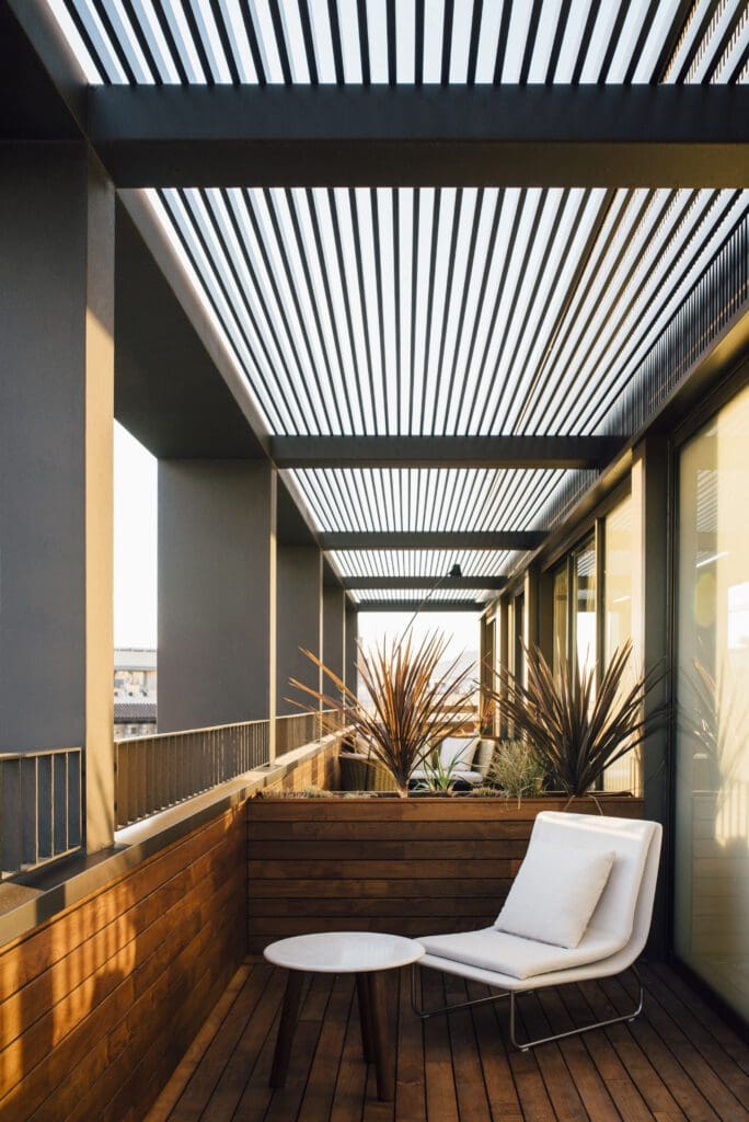 A modern balcony with wooden flooring, a white cushioned lounger, a small round table, potted plants, and a slatted pergola overhead.