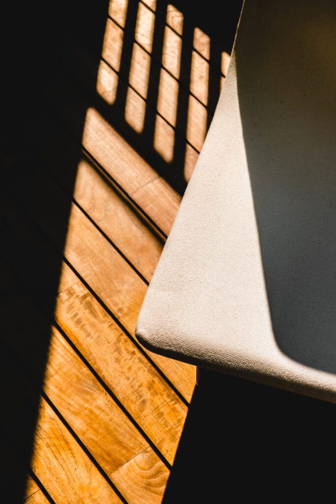Sunlight casts shadows from railing onto wooden floor next to the edge of a light-coloured fabric chair.