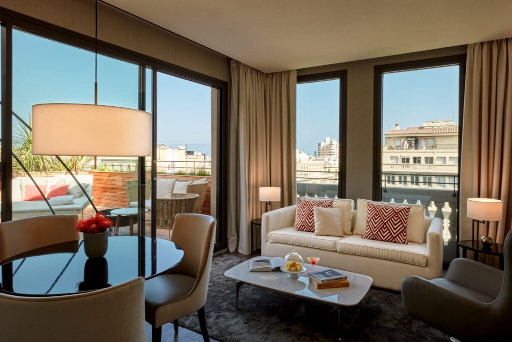 Modern living room with large windows showcasing a city view, a white sofa with patterned cushions, a round dining table, and a small outdoor balcony.