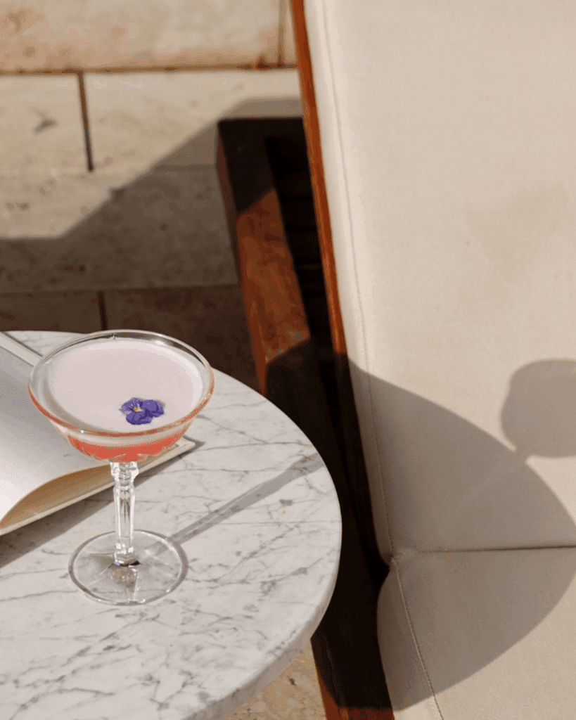 A coupe glass with a pink cocktail and a purple flower garnish sits on a round marble table next to an open book and a beige lounge chair.