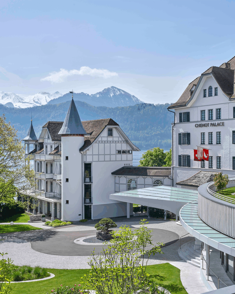 White hotel buildings with pointed towers and Swiss flags, set against a backdrop of mountains, trees, and a lake under a partly cloudy sky.