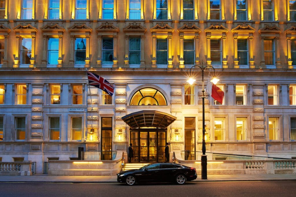 A black car is parked in front of an illuminated, ornate building with British and French flags displayed near the entrance at night.
