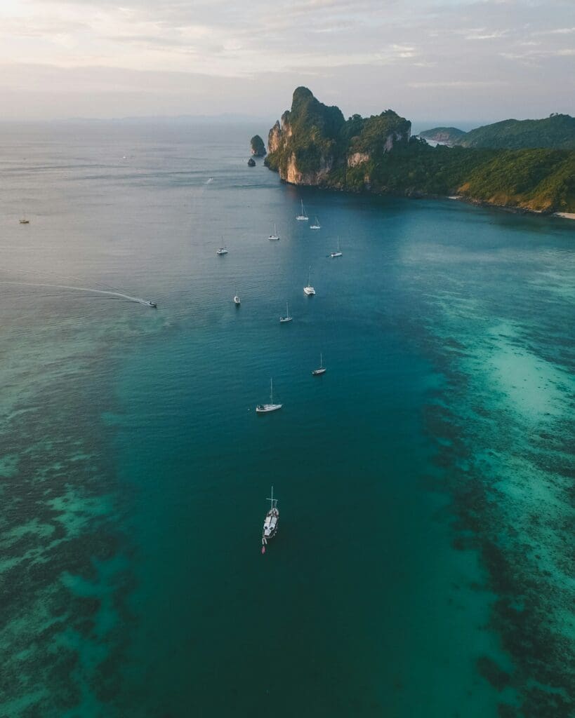 Aerial view of several sailing boats anchored in clear blue-green water near a lush, rocky island coastline under a partly cloudy sky.