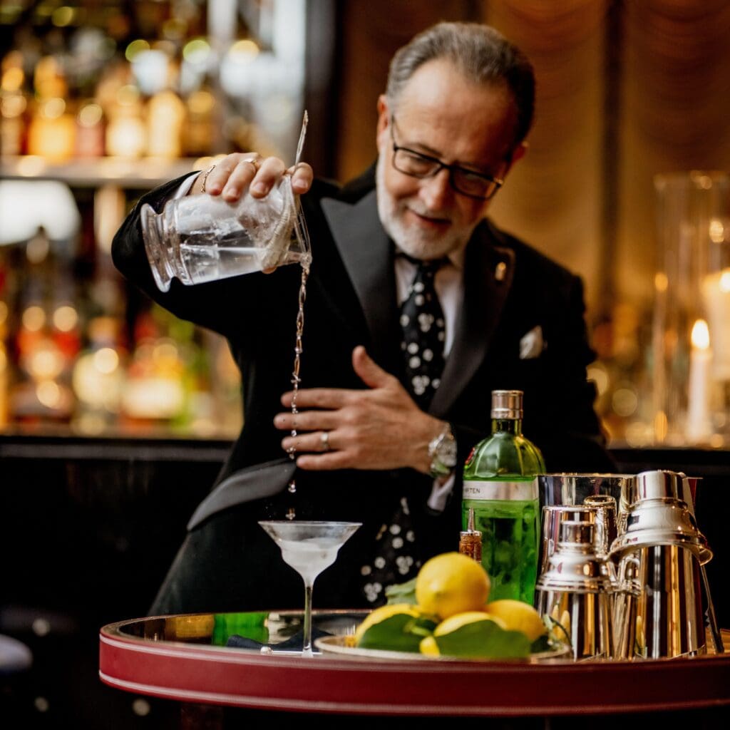 A barman in a suit pours a clear liquid from a mixing glass into a martini glass, with bottles, lemons, and cocktail shakers on the bar.