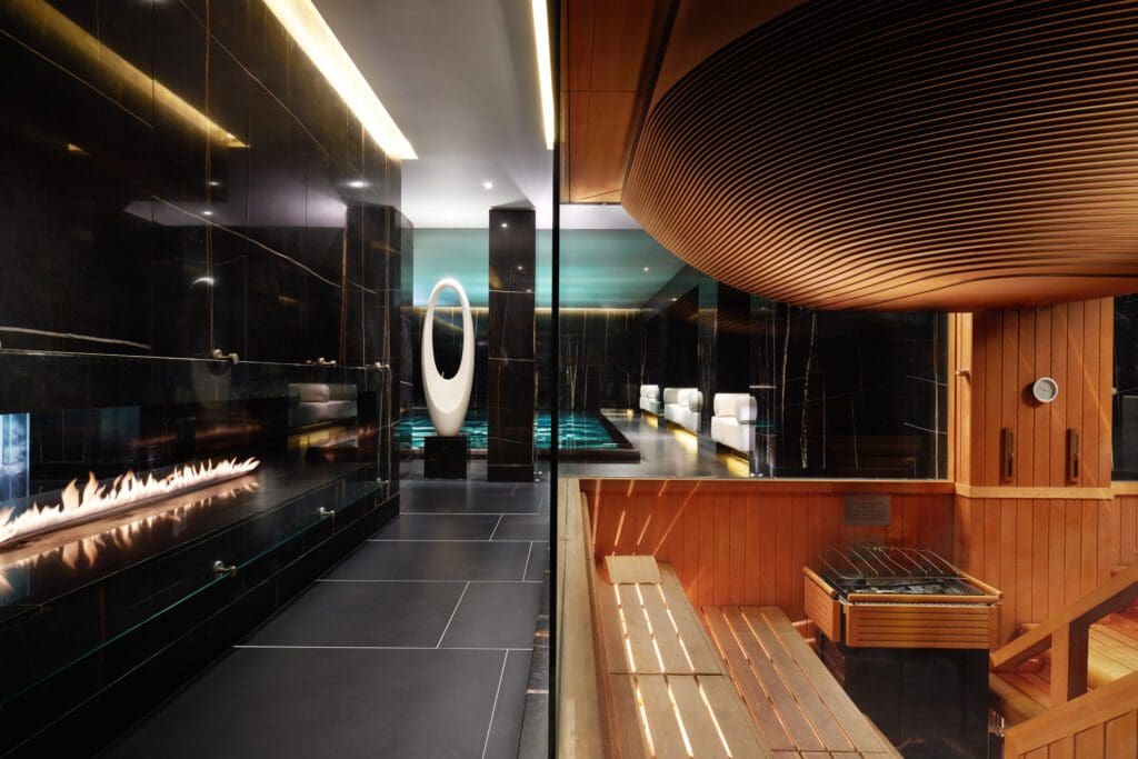 A modern spa interior with a wooden sauna on the right, a glass barrier, a long gas fireplace, and a sleek, dark-tiled relaxation area with ambient lighting.