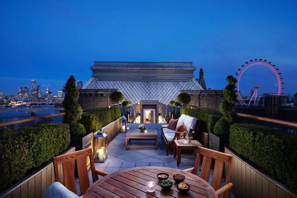 A rooftop terrace at dusk with seating, small tables, plants, and city views featuring the London Eye and skyline in the background.