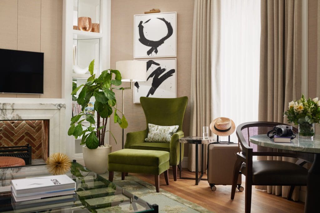 A lounge with a green armchair, potted plant, suitcase, hat, side table, abstract artwork, television, fireplace, and a round dining table with flowers. Natural light enters through curtains.