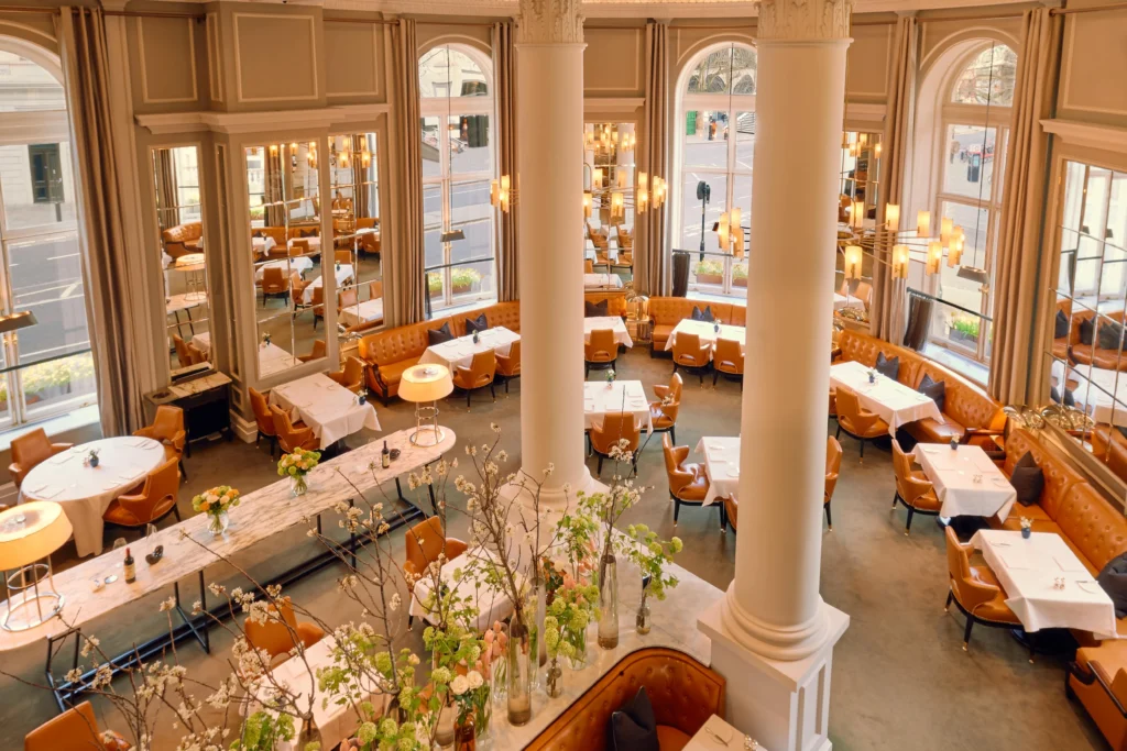 Spacious, elegant restaurant with tall arched windows, tan leather seating, white tablecloths, modern lighting, and decorative plants on tables.