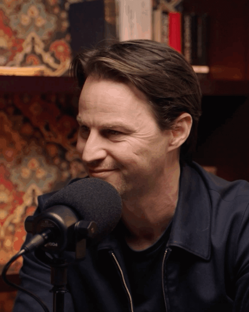 A man with brown hair sits in front of a microphone, smiling slightly, with books and patterned wallpaper in the background.