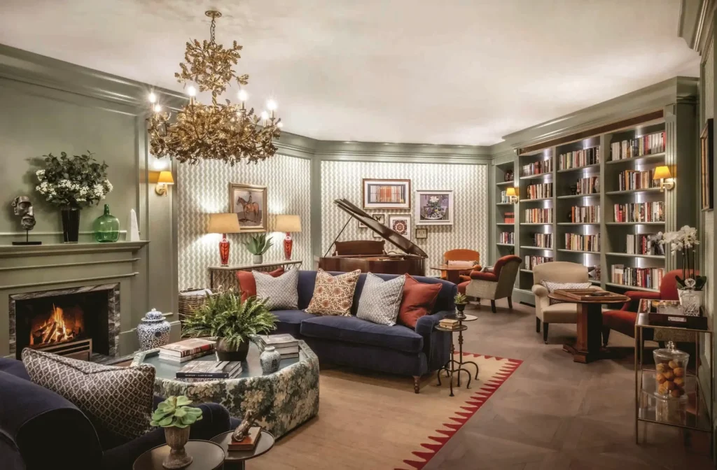 Elegant living room with a lit fireplace, blue sofas, bookcases, a piano, and a gold chandelier. Décor includes framed art, plants, and patterned wallpaper.