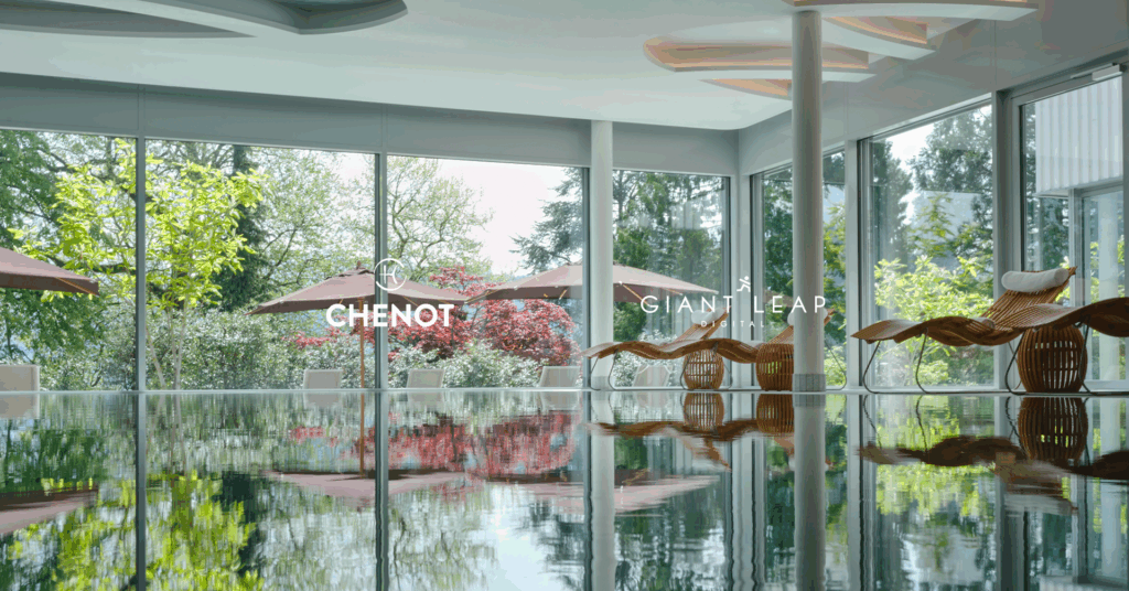 Indoor pool area with wicker loungers faces large windows overlooking a lush garden with trees, red bushes, and outdoor parasols. "Chenot" and "Giant Leap" logos appear on the glass.