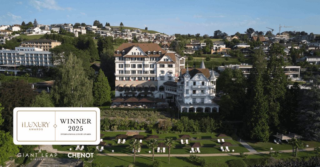 A large, elegant hotel with a white façade and brown roofs stands on landscaped grounds with lounge chairs; a "Winner 2025 iLuxury Awards" plaque is displayed in the foreground.